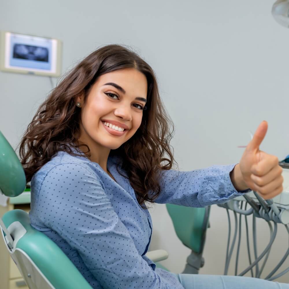 A woman in a dental chair smiles and gives a thumbs up